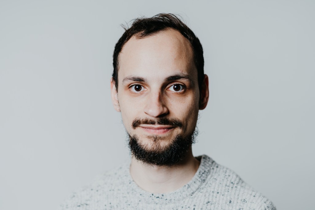Ein junger Mann mit Bart und grauem Pullover schaut in die Kamera