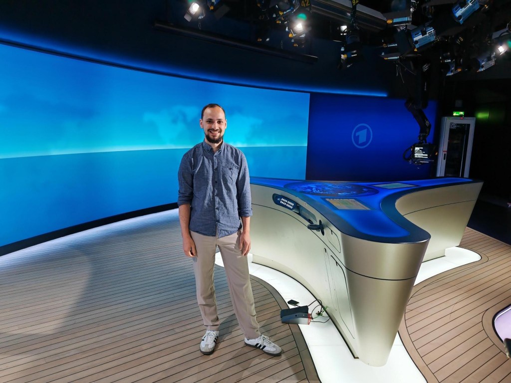 Ein junger Mann steht neben dem Moderationstisch im Tagesschau-Studio.